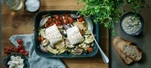 Baked Norwegian cod fillets with cherry tomatoes, olives, red onion and feta in an oven dish.